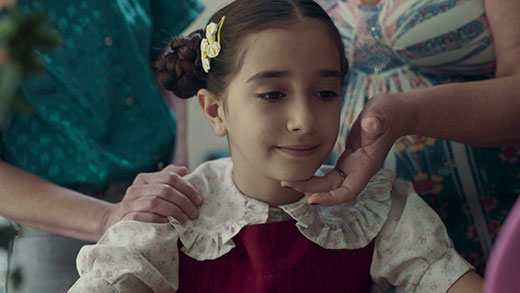 Uma menina com um vestido de gola de babados abre um sorriso desanimado. Ela tem o cabelo castanho escuro preso em coques ao lado da cabeça. Atrás dela, duas mulheres adultas lhe tocam no ombro e no queixo.