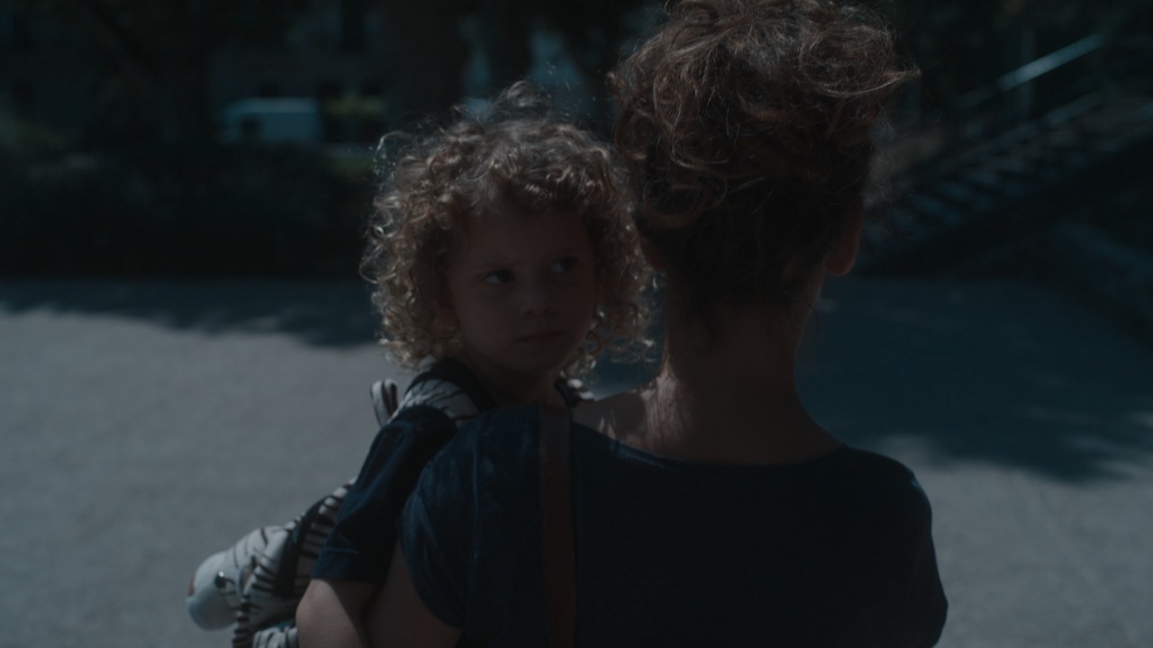 Mujer de espaldas caminando por la ciudad con un niño pequeño a upa. La mujer y la niña tienen el pelo rizado y claro. La niña lleva una mochila en formato de animal.