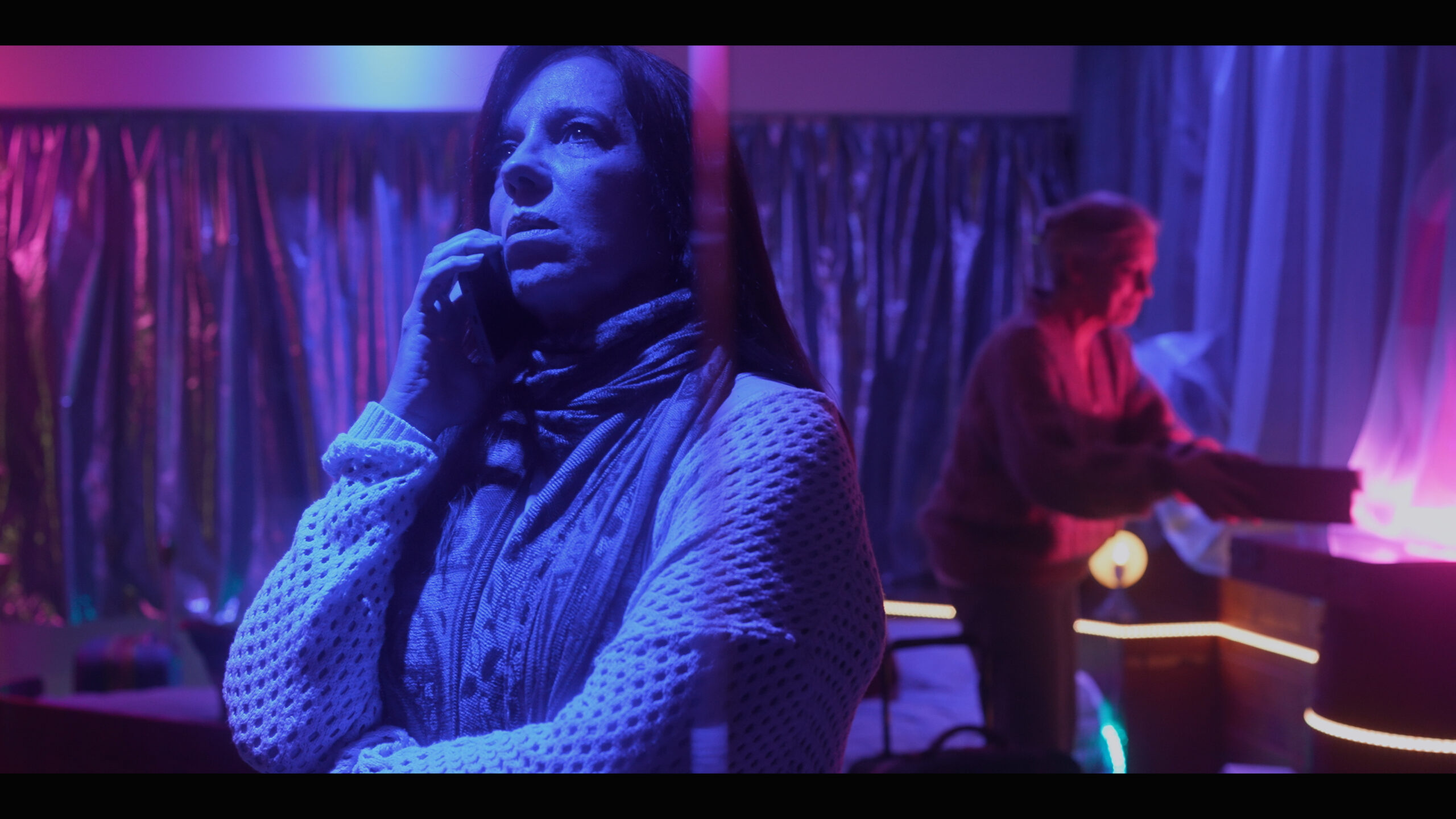 Una mujer de aproximadamente 65 años habla al teléfono. Ella está en una habitación, al fondo se ve a otra mujer, un poco más grande, con una pequeña caja en las manos. Todo está teñido con luces violáceas y magentas.