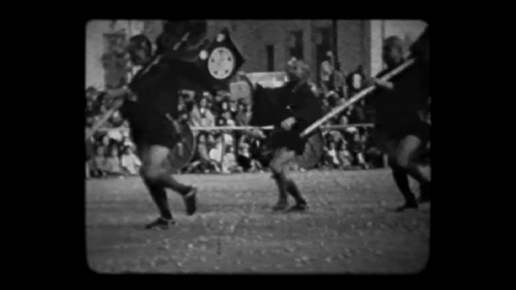El fotograma de una película antigua. La imagen está rodeada de márgenes negros y tiene las cuatro esquinas redondeadas. Imagen en blanco y negro. Fuera de foco, tres hombres asiáticos vestidos de negro caminan cargando objetos ceremoniales. Llevan pantalones cortos, están rapados al ras y al fondo una multitud los mira pasar.