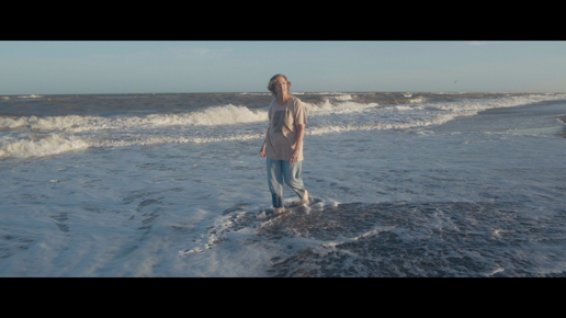 Mujer mayor, vestida con una camiseta clara y pantalones azules, está de pie dentro del mar, con el agua a la altura de los pies. Al fondo, se ven las olas y la extensión del océano.