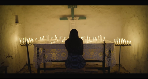 Mujer está sentada de espaldas frente a un altar cubierto con un tejido blanco, rodeado de decenas de velas encendidas. Sobre el altar, hay un crucifijo en la pared.
